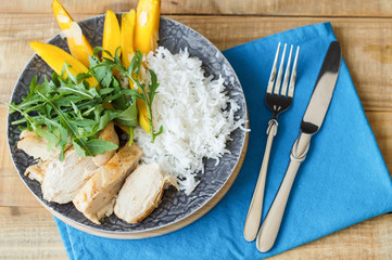 Plate with healthy food: white rice, chicken breast, arugula, spinach and mango fruit served with on blue napkin with fork and knife