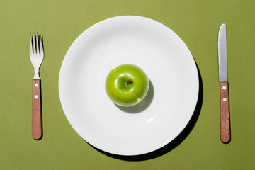 Top view of green apple on white plate with knife and fork on green background