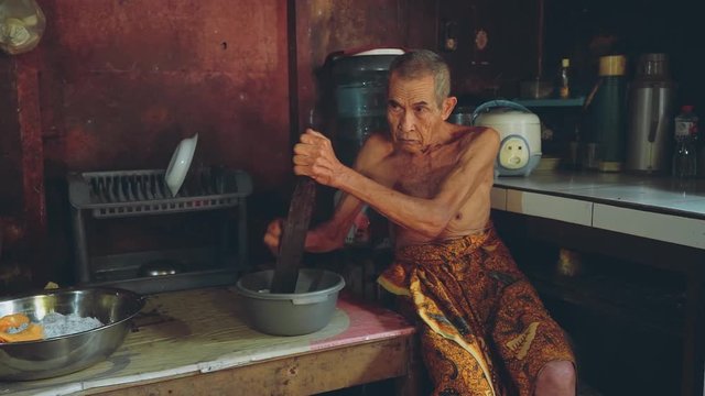 Balinese old man grating coconut in village kitchen, pan left