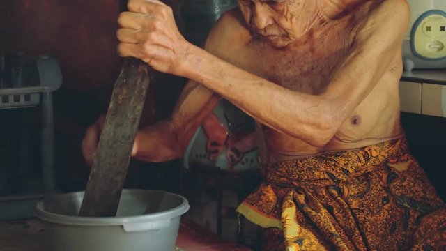 Balinese old man grating coconut in village kitchen, tilt down