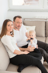 Happy family of three  is enjoying at home. Parents and their beautiful baby girl sitting on the sofa and looking at the camera. Family values. Leisure together.