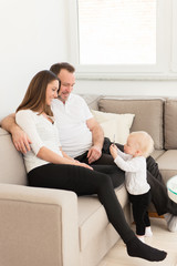 Obraz premium Happy family of three is enjoying at home. Parents sitting on the sofa and looking at their beautiful baby girl playing with cell phone. Family values. Leisure together.