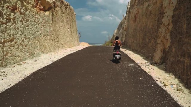 Woman riding black scooter in canyon, then beach road, view from back