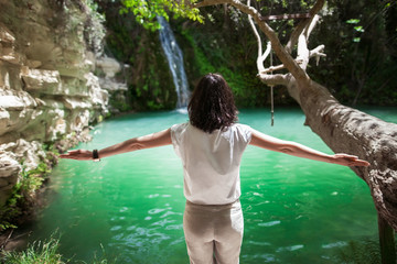 Back view of young woman with hands up enjoy waterfall on beautiful lake