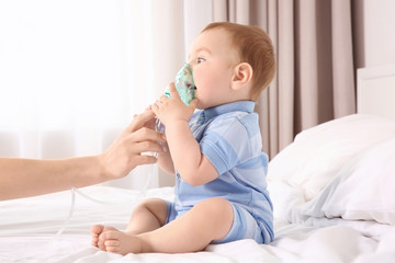 Mother helping her cute little baby with nebulizer while sitting on bed at home. Health care concept