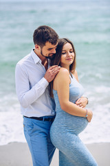 Obraz premium Courageous and handsome man in a white shirt and blue trousers hugs her beautiful pregnant wife in a blue dress against the background of the sea.