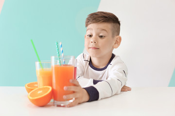 Cute little boy making long arm for glass of juice while sitting at table, on color background