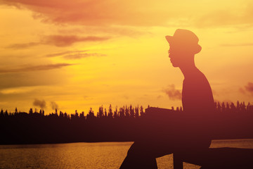 Fototapeta premium Silhouette of young man reading a book on the riverside park in free time.