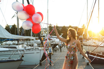 Woman in beautiful dress with a lot of colorful balloons on the yacht pier