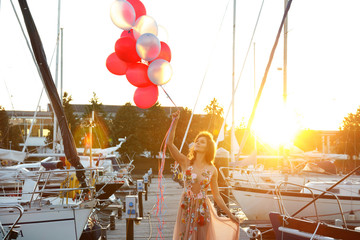 Woman in beautiful dress with a lot of colorful balloons on the yacht pier