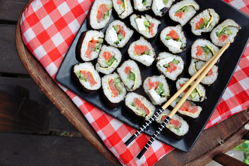 Sushi on a black square dish with chopsticks on a checkered red cloth, top view.