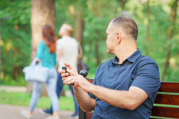adult man is looking at young couple sitting with phone at bench of summer park
