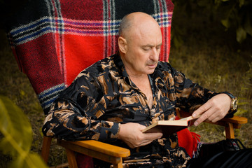 Fototapeta premium Mature stylish man read a book in garden in plaid armchair at autumn day
