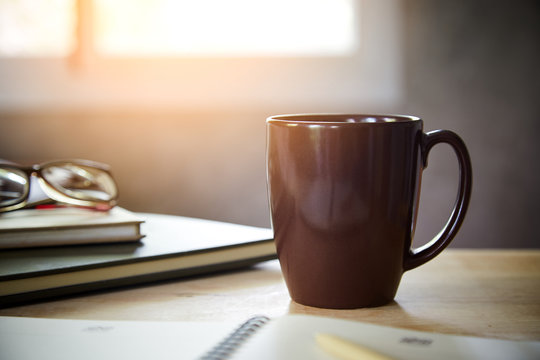 Morning coffee on the desk with sunlight from window, 
Coffee mug on the table with the blurred of notebook and glasses