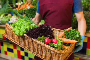 A friendly male seller posing with a huge basket of vegetables. Fresh vegetables in the background.