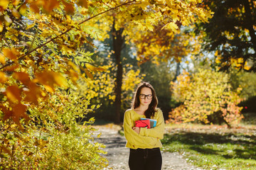 Portrait of pretty student girl with books in a autumn park, adult schoolgirl enjoying education, beautiful female wearing with yellow sweater , scarf and glasses, back to school