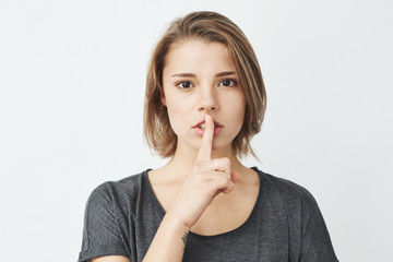 Young beautiful girl in grey t-shirt looking at camera showing keep silence over white background.