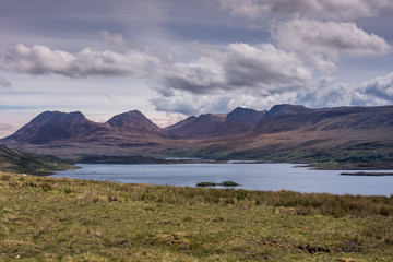 Assynt Peninsula, Scotland - June 7, 2012: Looking over Atlantic Ocean inlet towards brown mountain range under heavy sky with  white clouds. Green pasture foreground. Shot from near Loch Bad A Ghaill