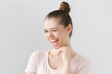Closeup shot of beautiful teenage girl isolated on grey background, looking caught by hilarious joke, laughing with open mouth, winking, pressing chin with fingers, showing excitement and happiness.
