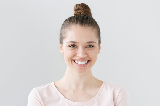 Indoor portrait of young attractive green-eyed female without makeup isolated on grey background in daylight smiling widely and happily, looking open and satisfied with communication and life.