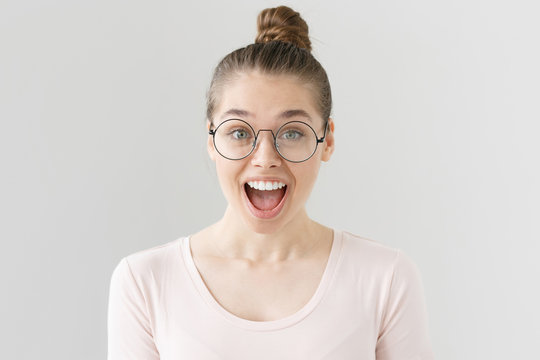 Indoor Shot Of Attractive Teenage Girl With Brown Hair Tied In Bun Isolated On Grey Background, Smiling Happily With Mouth Open In Surprise, Anticipating Cool Prospects In Future With Pleasure.