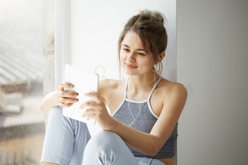 Fototapeta premium Portrait of young teenage cheerful girl in headphones smiling looking at tablet surfing web browsing internet sitting near window over white wall.