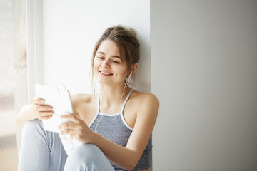 Portrait of young teenage cheerful girl in headphones smiling looking at tablet surfing web browsing internet sitting near window over white wall.