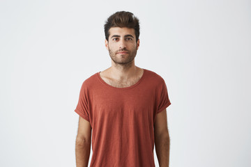 Portrait of stylish bearded guy with trendy haircut wearing casual red T-shirt looking with his brown eyes at the camera. Young handsome man having pleased look. People and emotions concept.