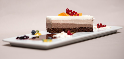 Fancy triple mousse chocolate dessert, placed on a white plate, decorated with forest fruits and chocolate sauce, white plate, light brown background