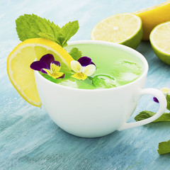 Cold refreshing drink with mint syrup, ice cubes, mint, lemon, edible flowers of a garden viola on a light turquoise summer background in a white ceramic cup. Selective focus. Top View.