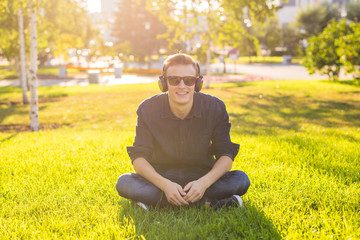 Naklejka na ściany i meble Portrait of a relaxed young man sitting on grass in park and listening to music on headphones.