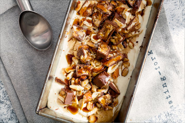 Snickers ice cream with caramel,  fried peanuts and hazelnuts in metallic baking tray on rustic background. Gourmet summer treat concept. Composition with copy space.