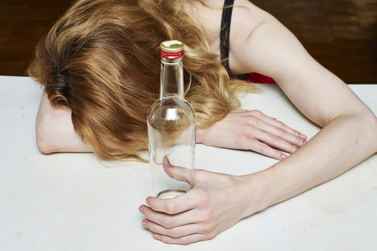Drunk woman holding an bottle vodka and sleeping with her head on the table