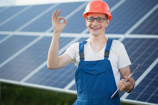 Worker on solar power station. Young man smailing, showing ok with his fingers, solar panels behind him.
