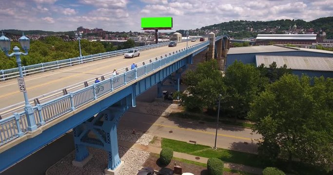 A slow reverse aerial establishing shot of traffic and bicyclists on the 31st Street Bridge in the Lawrenceville area of Pittsburgh. Green screen billboard for customization.  	