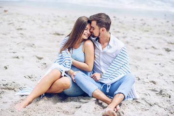 Fototapeta na wymiar Beautiful pregnant woman and her respectable man leaning against each other and covered with a white striped plaid sitting on the seashore. Lovely young couple.