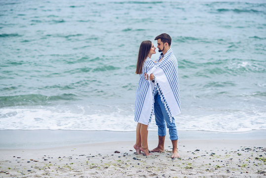 Beautiful pregnant woman and her respectable man leaning against each other and coveredwith a white striped plaid on the seashore. Lovely young couple, future parents.