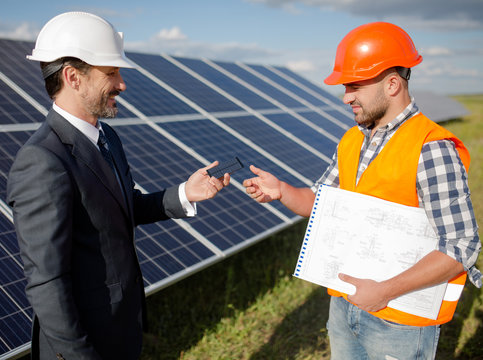 Business client stretching out photovoltaic item to foreman. Solar station in the open air, client and worker discussing technical details.