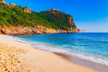 Cleopatra beach on sea coast with green rocks in Alanya peninsula, Antalya district, Turkey. Beautiful sunny landscape for tourism with clear water and sand. Alanya Castle on the cliff