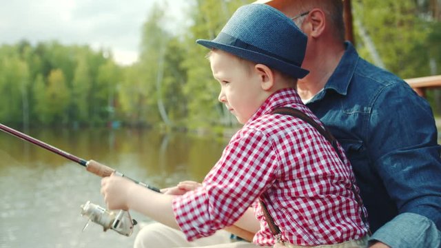 Little boy is turning fishing reel on fishing with his grandfather at the lake