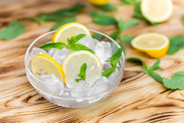 Fresh sliced lemon, bright green mint leaves and frozen ice cubes in a transparent bowl on a wooden table