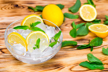 Fresh sliced lemon, bright green mint leaves and frozen ice cubes in a transparent bowl on a wooden table