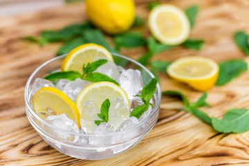 Fresh sliced lemon, bright green mint leaves and frozen ice cubes in a transparent bowl on a wooden table