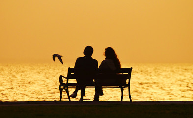 Fototapeta na wymiar Young couple sitting on a bench near seashore at sunset, silhouette. A bird flying on the sea. 