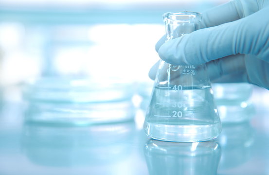 scientist hand in rubber glove holding clear flask with blur petri dish background in medical research science laboratory