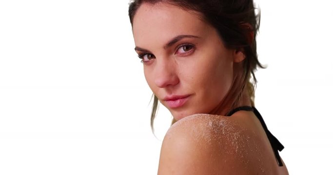 Close up of attractive Caucasian millennial looking over shoulder at camera on white background with copyspace. Closeup of beautiful young woman in string bikini with sand on shoulder in studio. 4k 