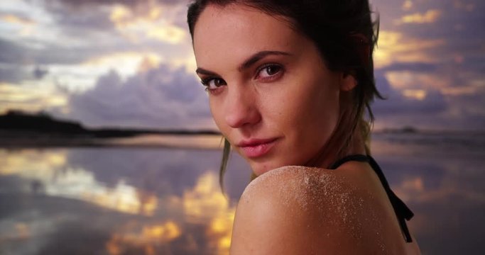 Close up of Caucasian millennial standing on the beach at sunset, looking at camera with flirty expression. Closeup of beautiful young woman in string bikini looking over shoulder at dusk. 4k 