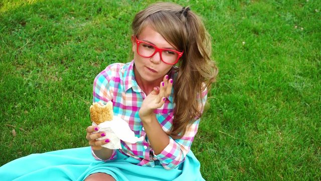 Young attractive girl eating a sandwich in an outdoor park. Hipster girl in a plaid shirt and glasses. Student at the break. Fast food.