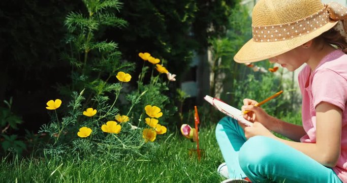 Girl aged 8 looking drowing in notebook and sitting on the grass in the garden at sunny day and smiling at camera