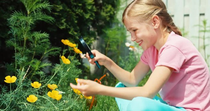 Girl aged 8 looking through magnifying glass at flower. Kid sitting on the grass in the garden at sunny day and smiling at camera